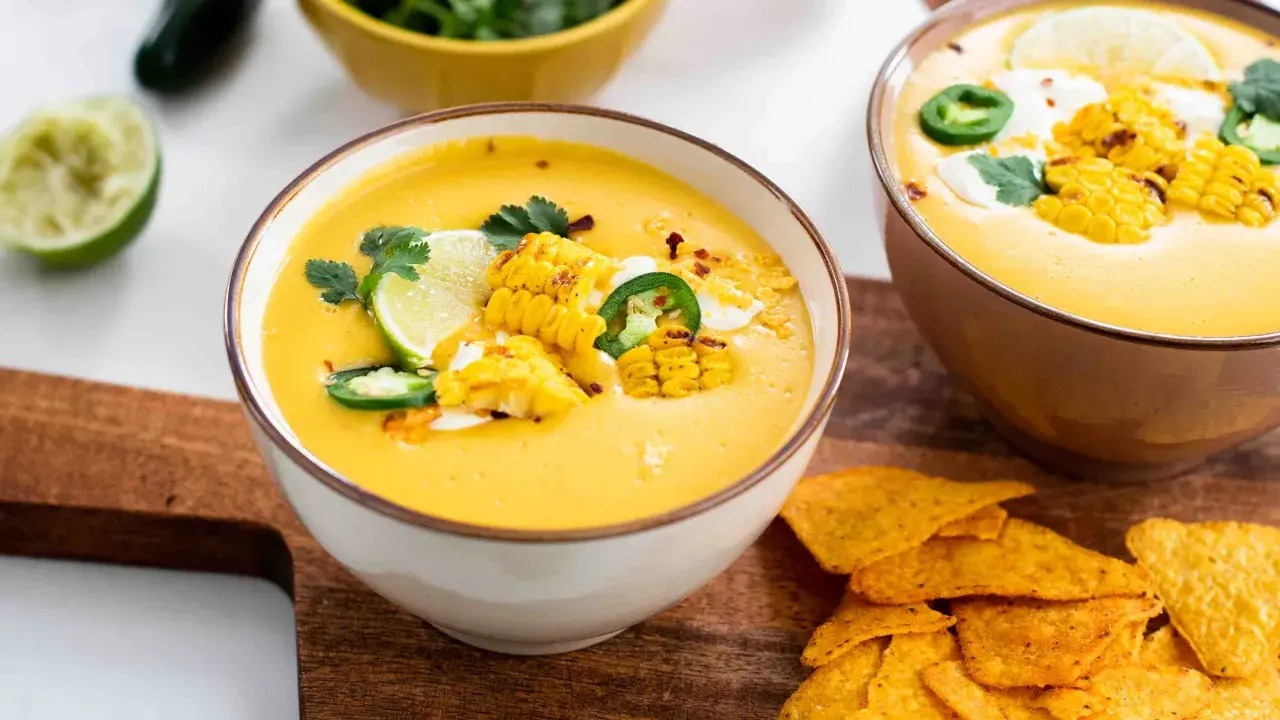Zwei Schüsseln mit cremiger Mais-Suppe, garniert mit Maiskolben, Limette und Jalapeños, daneben Tortilla-Chips.
